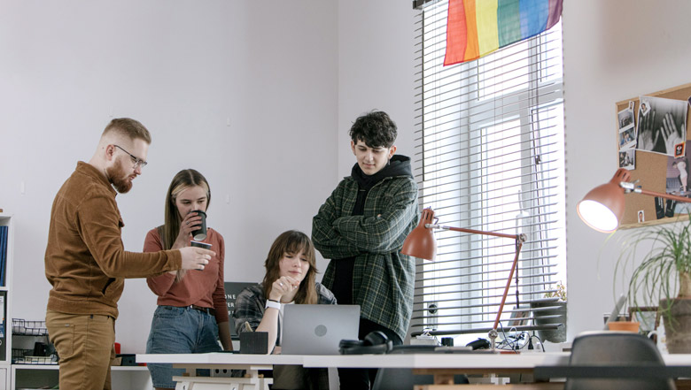 young people gathered around a laptop in an office with a pride flag behind them